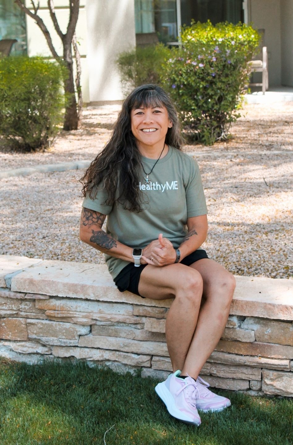 Woman in "Healthy ME" shirt sits on wall.